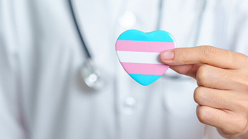 Herz mit Farben der trans* Flagge vor einer Person in einem medizinischen Kittel mit Stethoskop